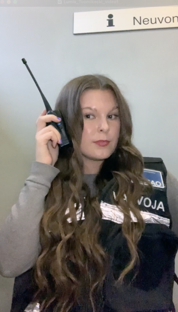 Henkilö, jolla on pitkä, ruskea tukka, pitää radiopuhelinta korvallaan. Hänellä on päällään liivi, jossa lukee 'Valvoja'.