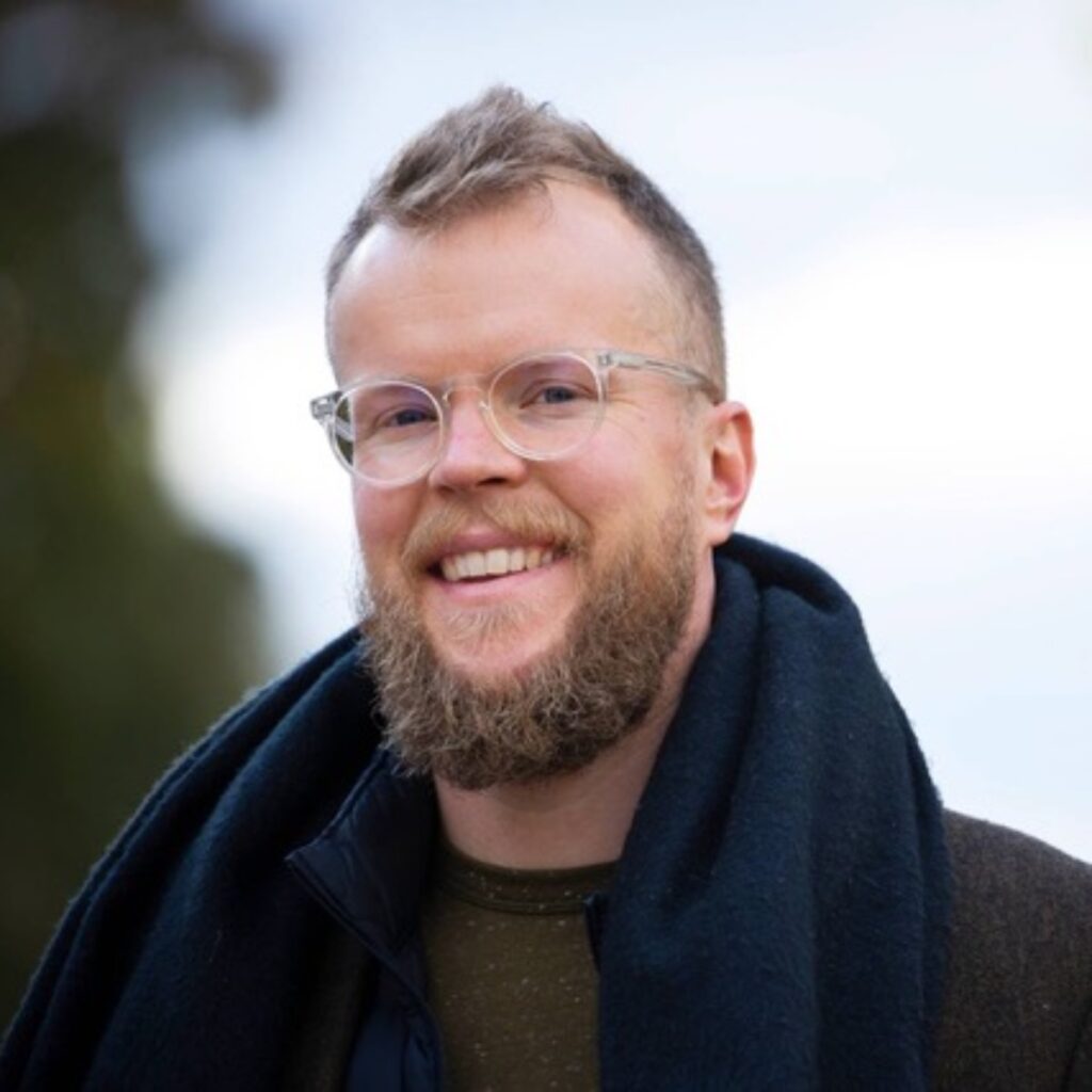 Mies, jolla on parta ja lasit, hymyilee kameralle ulkona.