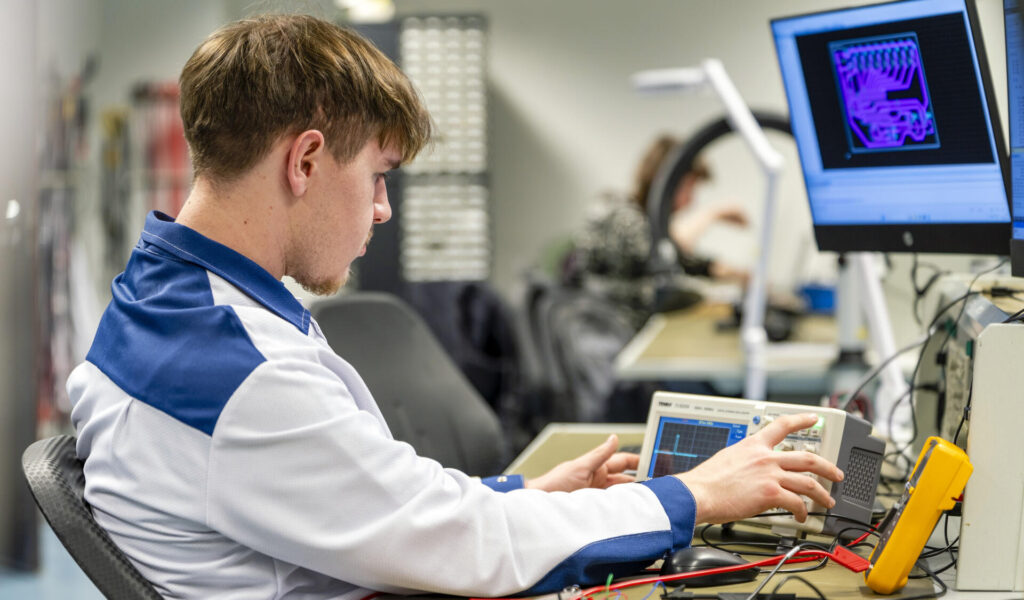 Nuori mies istuu työpöydän ääressä laboratoriotakissa ja käyttää oskilloskooppia. Näytöllä näkyy teknisiä piirustuksia. Hän on keskittynyt elektroniikan säätöihin.