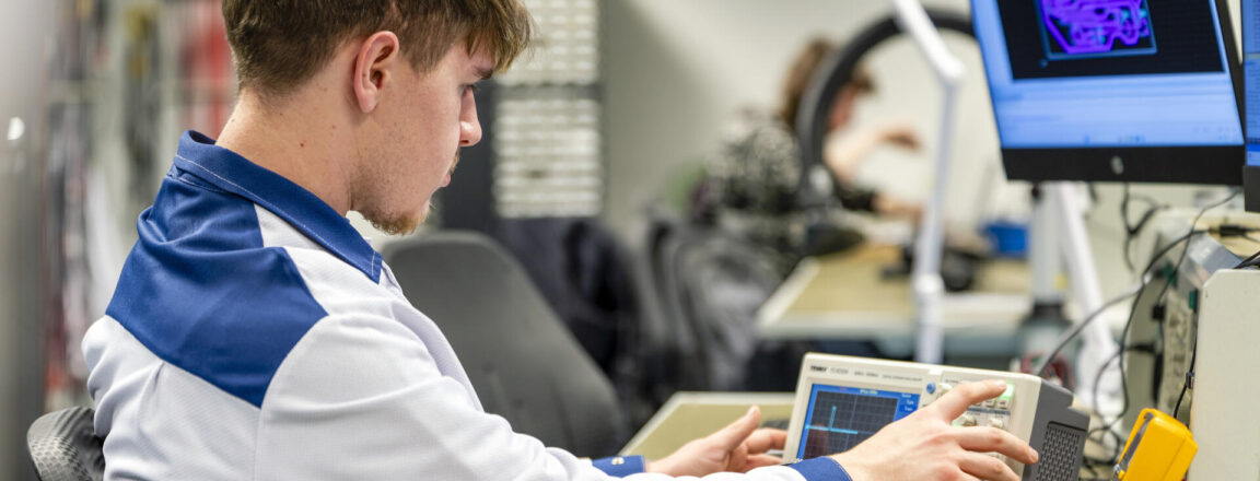 Nuori mies istuu työpöydän ääressä laboratoriotakissa ja käyttää oskilloskooppia. Näytöllä näkyy teknisiä piirustuksia. Hän on keskittynyt elektroniikan säätöihin.