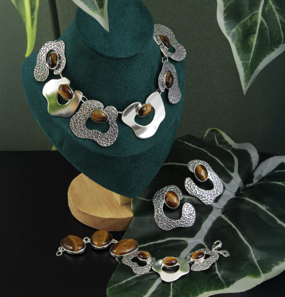 Koruja esillä vihreällä alustalla: kaulakoru ja korvakorut, joissa on ruskeita kiviä ja hopeanvärisiä kuvioita. Taustana vihreitä lehtiä.