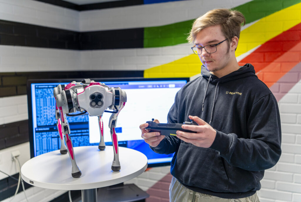 Henkilö ohjaa robottikoiraa kaukosäätimellä. Robottikoira on pöydällä ja taustalla on tietokoneen näyttö, jossa on teknistä tietoa. Huoneen seinä on koristeltu värikkäin viivoin.