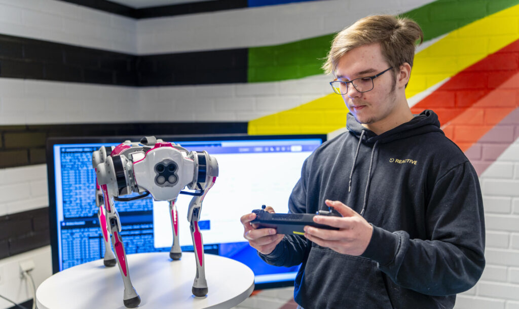 Henkilö seisoo sisätiloissa ja pitää tablettia kädessään. Hänen edessään on pöydällä robottikoira. Taustalla on iso näyttö ja värikäs seinä.