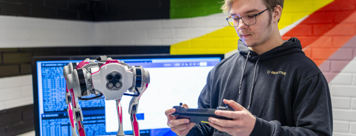 Henkilö seisoo sisätiloissa ja pitää tablettia kädessään. Hänen edessään on pöydällä robottikoira. Taustalla on iso näyttö ja värikäs seinä.