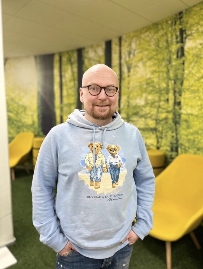 Mies seisoo sisätilassa, taustalla on metsämaisema. Hänellä on päällään vaaleansininen huppari, jossa on nallekarhujen kuva.