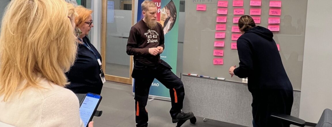 Neljä henkilöä seisoo huoneessa valkotaulun edessä, jossa on paljon vaaleanpunaisia post-it-lappuja. Yksi henkilö kirjoittaa taululle, kun muut katsovat.