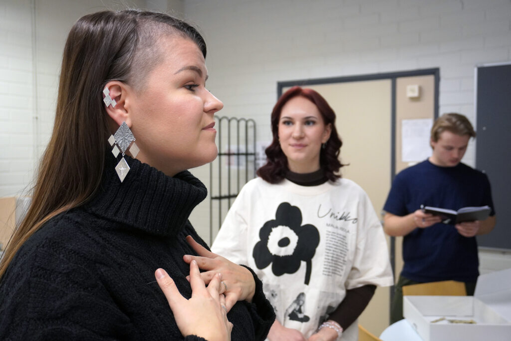 Kolme henkilöä huoneessa: nainen vasemmalla mustassa villapuserossa ja suuret korvakorut korvissaan, taustalla nainen valkoisessa Unikko-paidassa katsoo toista kohti, ja mies taustalla oikealla katsoo muistikirjaa kynä kädessään.