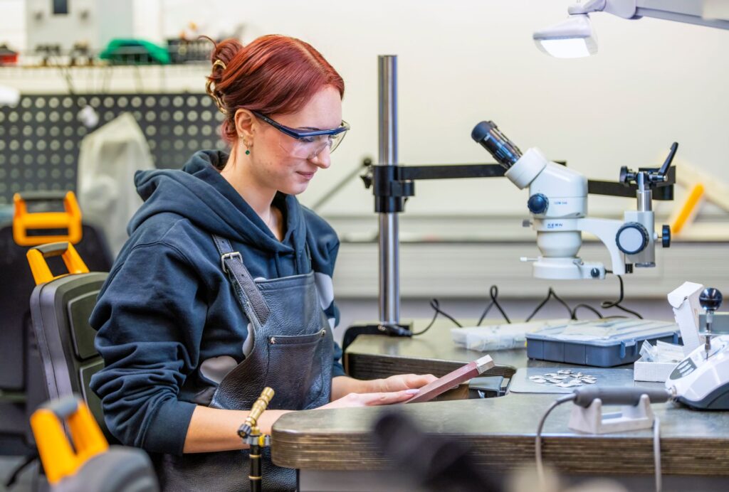 Henkilö istuu työpöydän ääressä työpajassa, jossa valmistetaan koruja. Hänellä on yllään huppari ja suojalasit. Pöydällä on metallisia korun osia ja mikroskooppi.