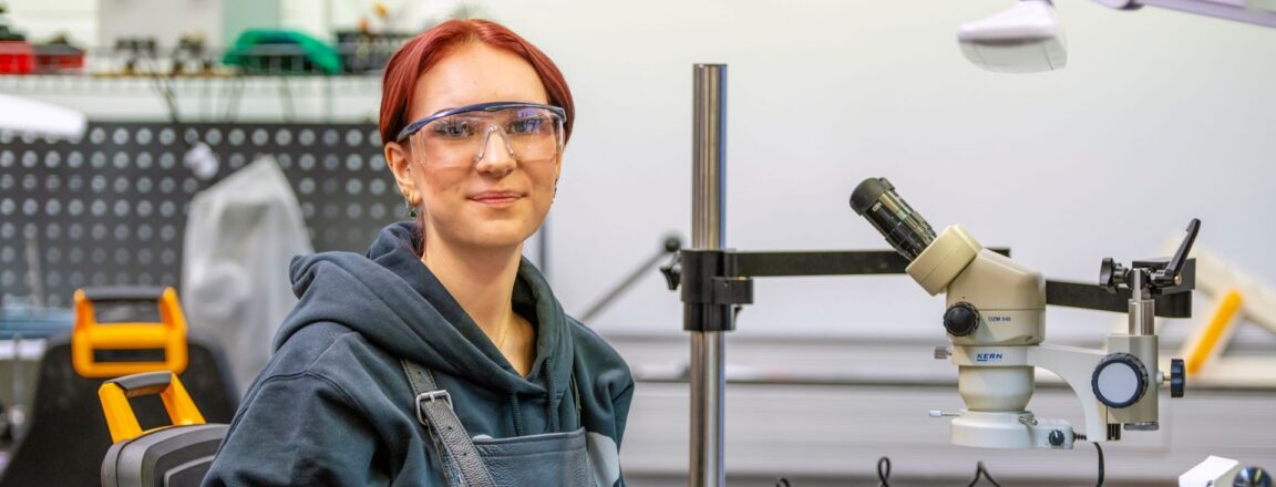 Henkilö istuu työpöydän ääressä työpajassa, jossa valmistetaan koruja. Pöydällä on metallisia korun osia ja mikroskooppi. Hänellä on yllään huppari ja suojalasit.