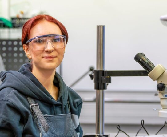 Henkilö istuu työpöydän ääressä työpajassa, jossa valmistetaan koruja. Pöydällä on metallisia korun osia ja mikroskooppi. Hänellä on yllään huppari ja suojalasit.