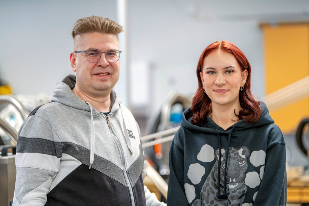 Mies ja nainen seisovat vierekkäin sisätiloissa. Mies on pukeutunut harmaa-mustaan huppariin ja nainen mustaan huppariin, jossa on leopardikuvio. Taustalla näkyy toimistolaitteita.