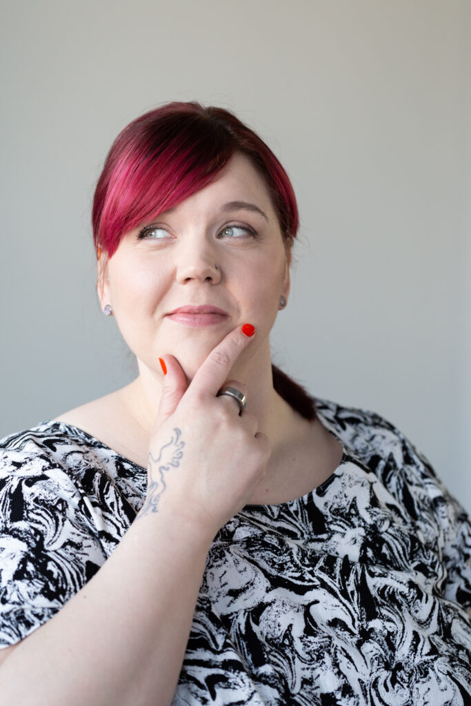 Punahiuksinen nainen katsoo hymyillen sivulle, käsi nostettuna leuan alle. Naisella on mustavalkokuvioinen paita ja kirkkaanpunaiset kynnet.