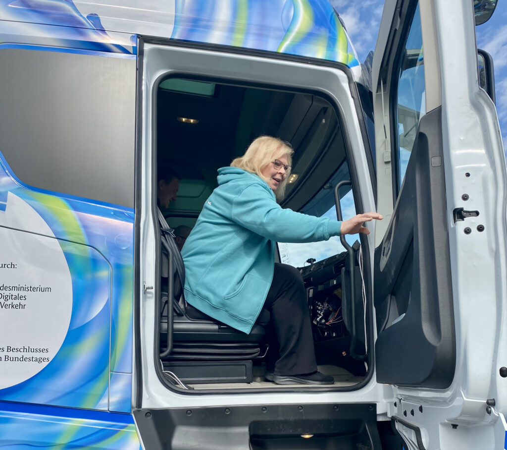 Henkilö sinisessä hupparissa nousee rekka-auton ohjaamoon. Rekan ovi on auki ja taustalla näkyy sinistä ja vihreää väritystä rekassa.