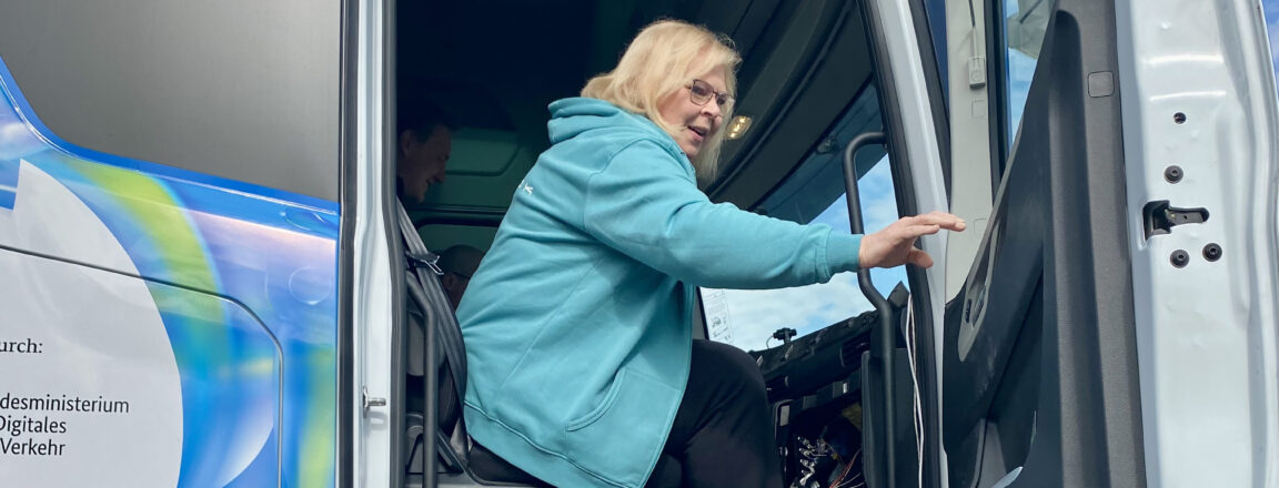 Henkilö sinisessä hupparissa nousee rekka-auton ohjaamoon. Rekan ovi on auki ja taustalla näkyy sinistä ja vihreää väritystä rekassa.