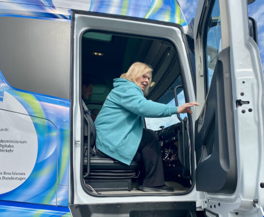 Henkilö sinisessä hupparissa nousee rekka-auton ohjaamoon. Rekan ovi on auki ja taustalla näkyy sinistä ja vihreää väritystä rekassa.