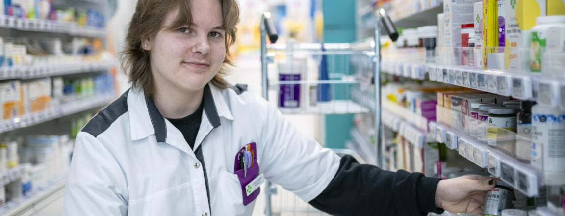 Henkilö on kyykyssä apteekissa tuotteiden keskellä ja hymyilee kameralle. Hänellä on valkoinen takki ja hän järjestelee hyllyllä olevia tuotteita.