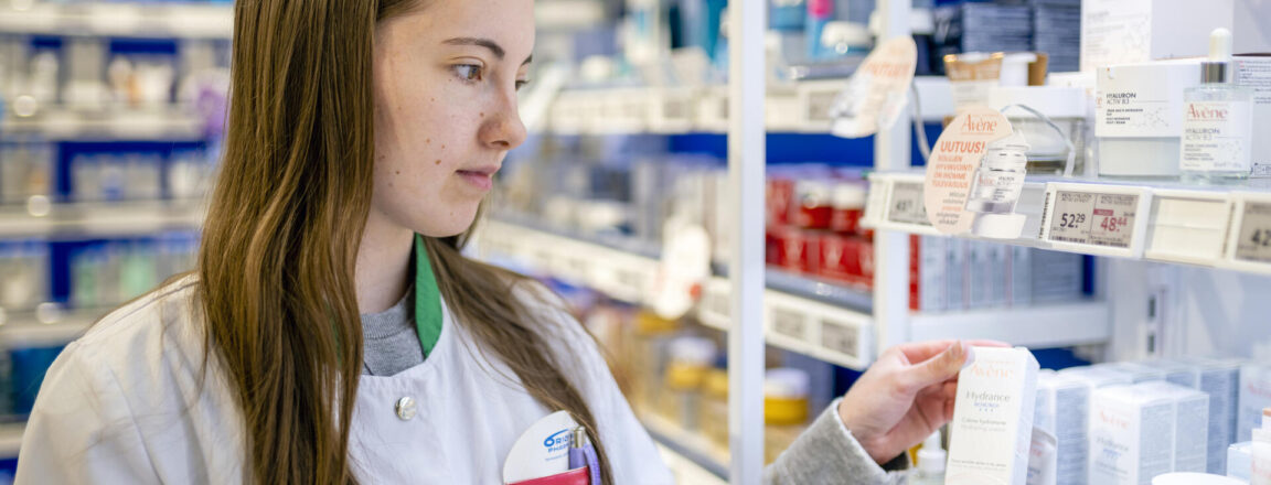 Nainen seisoo apteekissa ja tarkastelee hyllyllä olevia tuotteita. Hänellä on yllään valkoinen takki ja hän katsoo huolellisesti yhtä tuotepakettia kädessään.