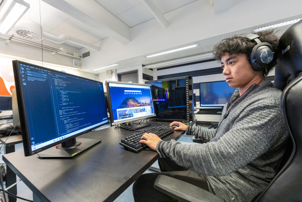 Henkilö istuu tietokoneen ääressä toimistoympäristössä, kahden näytön edessä. Henkilöllä on kuulokkeet ja hän näyttää keskittyvän ohjelmointitehtävään.