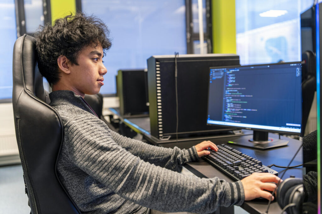 Nuori henkilö istuu tietokoneen ääressä koodaten. Hän on keskittynyt näyttöön, joka näyttää ohjelmointikoodia. Tietokoneet ovat rivissä toimistoympäristössä.