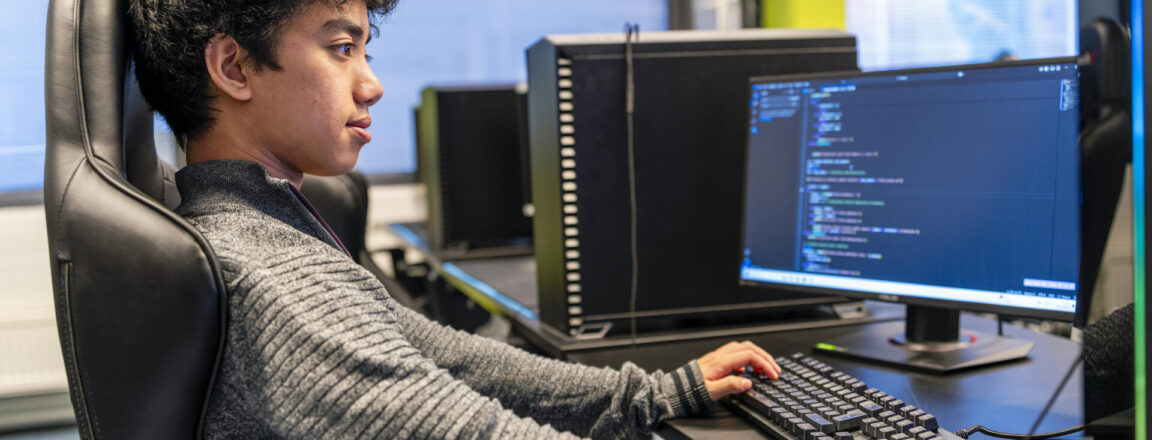 Nuori henkilö istuu tietokoneen ääressä koodaten. Hän on keskittynyt näyttöön, joka näyttää ohjelmointikoodia. Tietokoneet ovat rivissä toimistoympäristössä.