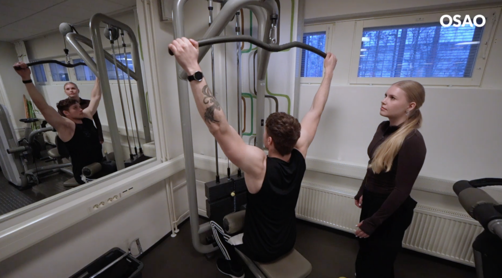 Mies käyttää kuntosalillla ylätaljalaitetta. Vieressä seisoo nainen, joka tarkkailee hänen suoritustaan. Huoneessa on peili ja ikkunat, joista näkyy talvinen maisema. Kuvassa näkyy myös OSAO-logo.