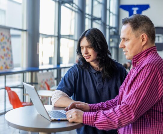 Kaksi henkilöä seisoo pöydän ääressä ja katsoo kannettavaa tietokonetta. Taustalla on ikkunoita ja värikkäitä tuoleja. Toinen henkilöistä on pukeutunut tummansiniseen puseroon ja toinen pinkkimustaan ruutupaitaan.