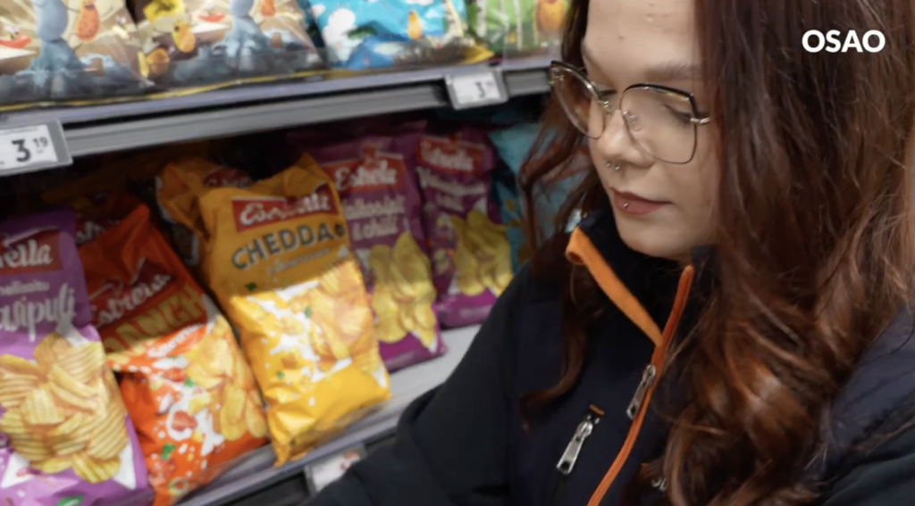 Nuori nainen laittamassa kaupassa sipsihyllylle lisää tuotteita. Hänellä on päällään kaupan työntekijän takki.