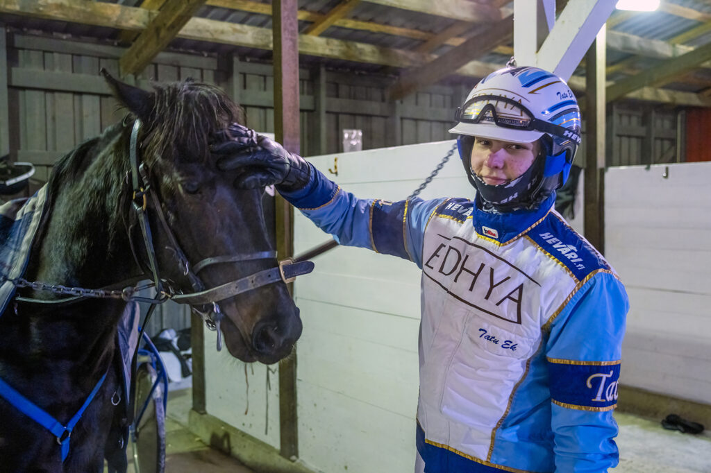 Ravikuski hymyilee ja silittää hevosta tallissa. Hänellä on päällään sinivalkoinen ajopuku ja kypärä, ja hevonen on valjaissa.