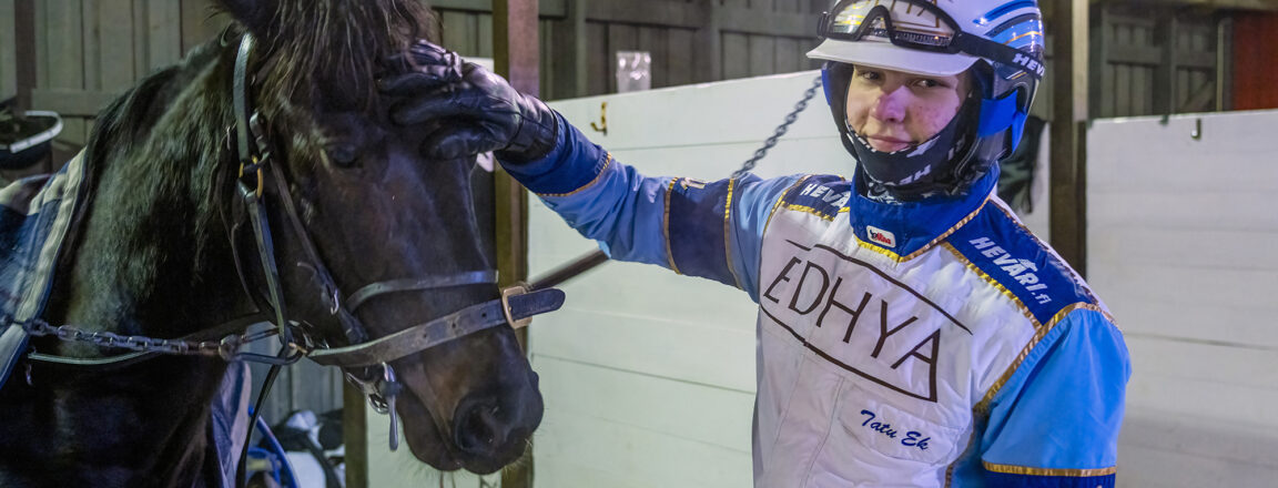 Ravikuski hymyilee ja silittää hevosta tallissa. Hänellä on päällään sinivalkoinen ajopuku ja kypärä, ja hevonen on valjaissa.