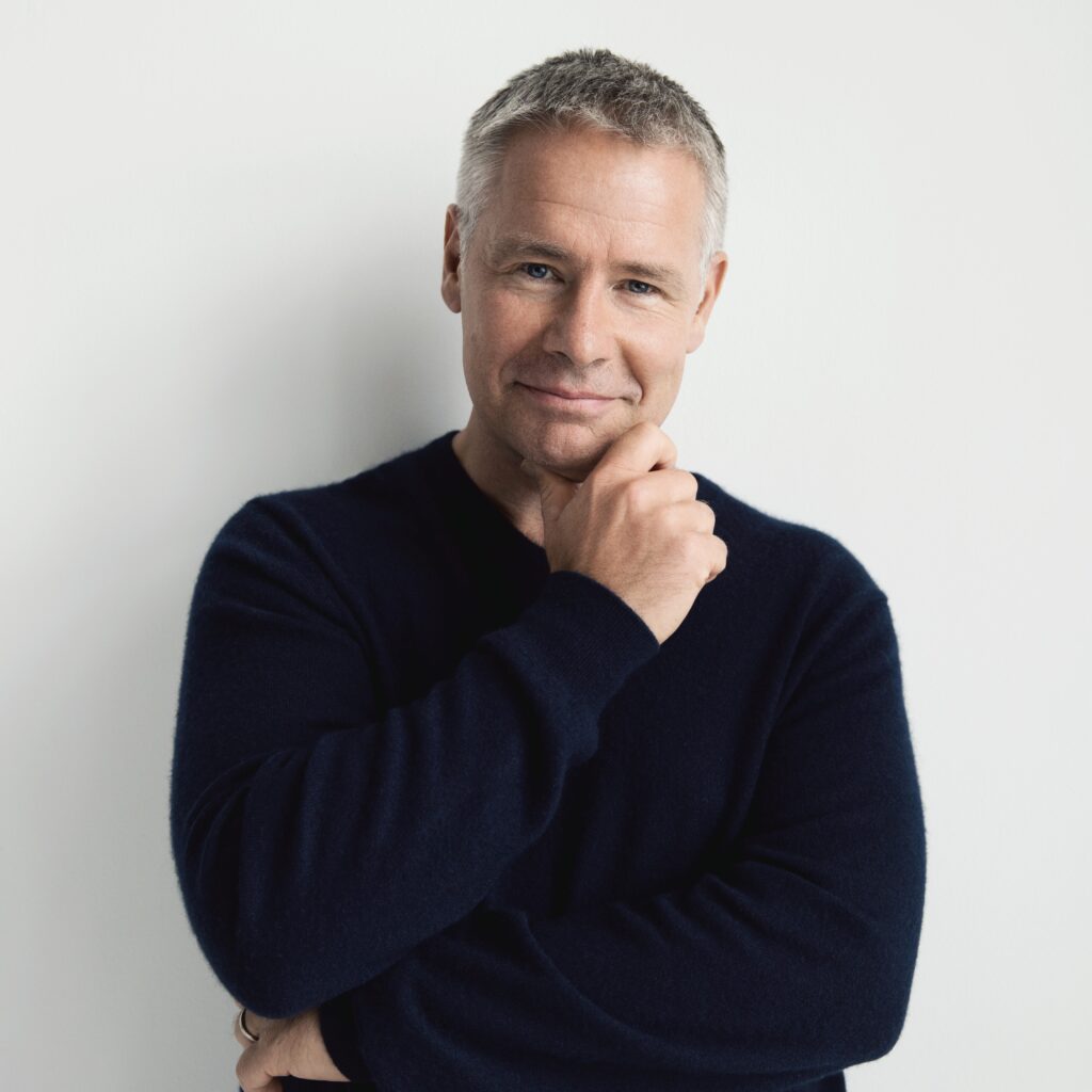 Mies tummassa neuleessa hymyilee käsi leuan alla valkoista taustaa vasten.