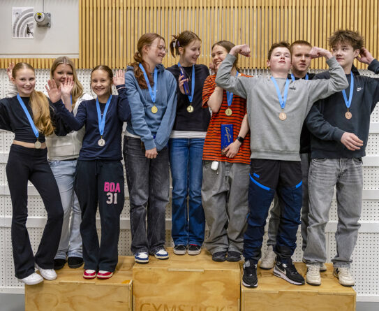 Ryhmä nuoria seisoo palkintokorokkeen edessä ja hymyilee. Heillä on kaulassaan mitaleita ja he näyttävät iloisilta. Taustalla on puinen seinä ja valkoinen tausta.