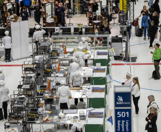 Leipomokilpailu sisätiloissa, jossa useita leipureita työskentelee pitkien työasemien ääressä. Taustalla on ihmisiä seuraamassa tapahtumaa. "SkillsFinland"-kyltti näkyy oikealla puolella. Kilpailijat ovat pukeutuneet valkoisiin työasuihin ja päähineisiin.