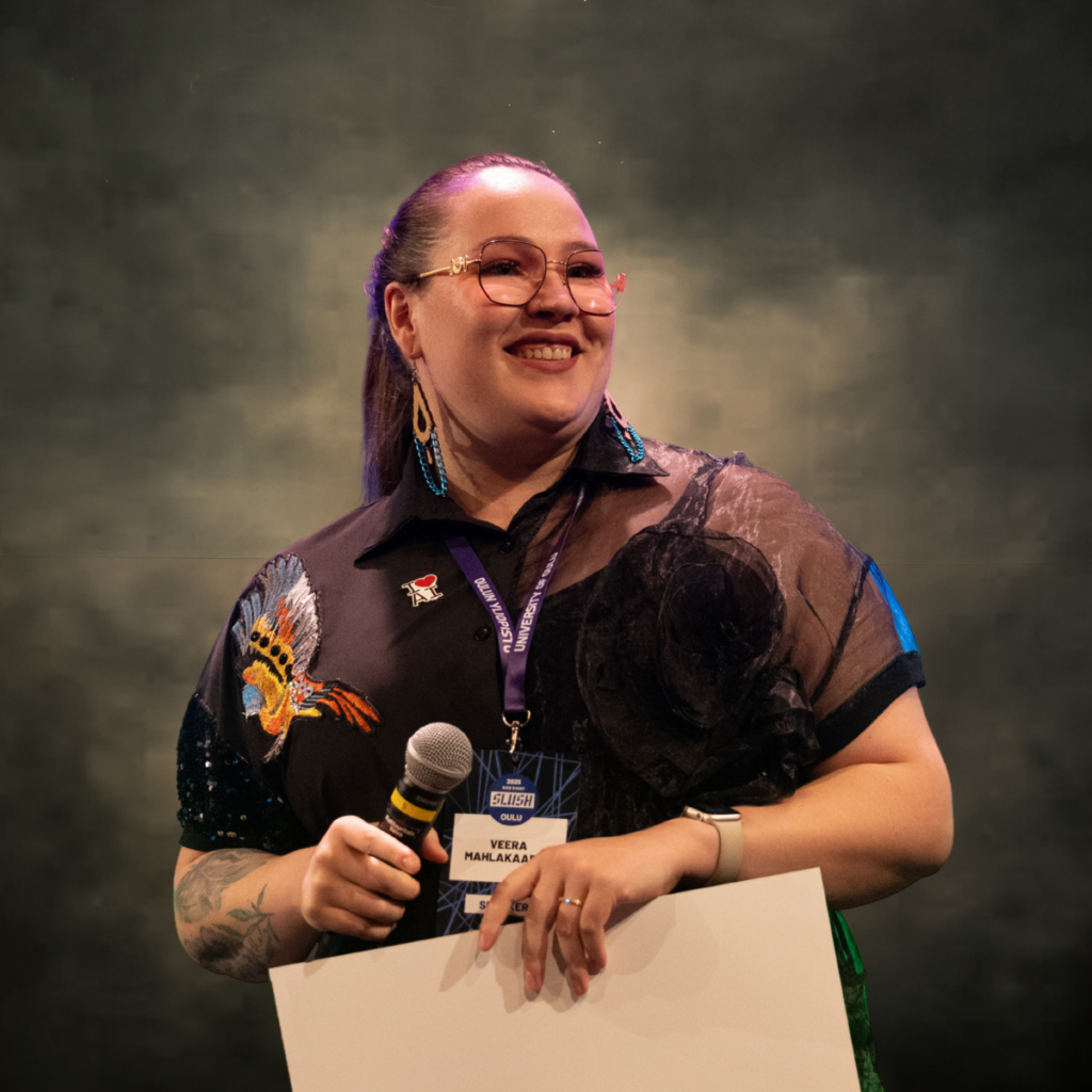 Henkilö seisoo lavalla pitäen toisessa kädessään mikrofonia ja toisessa paperia. Hänellä on silmälasit, isot korvakorut ja kuvioitu paita. Tausta on hämärästi valaistu.