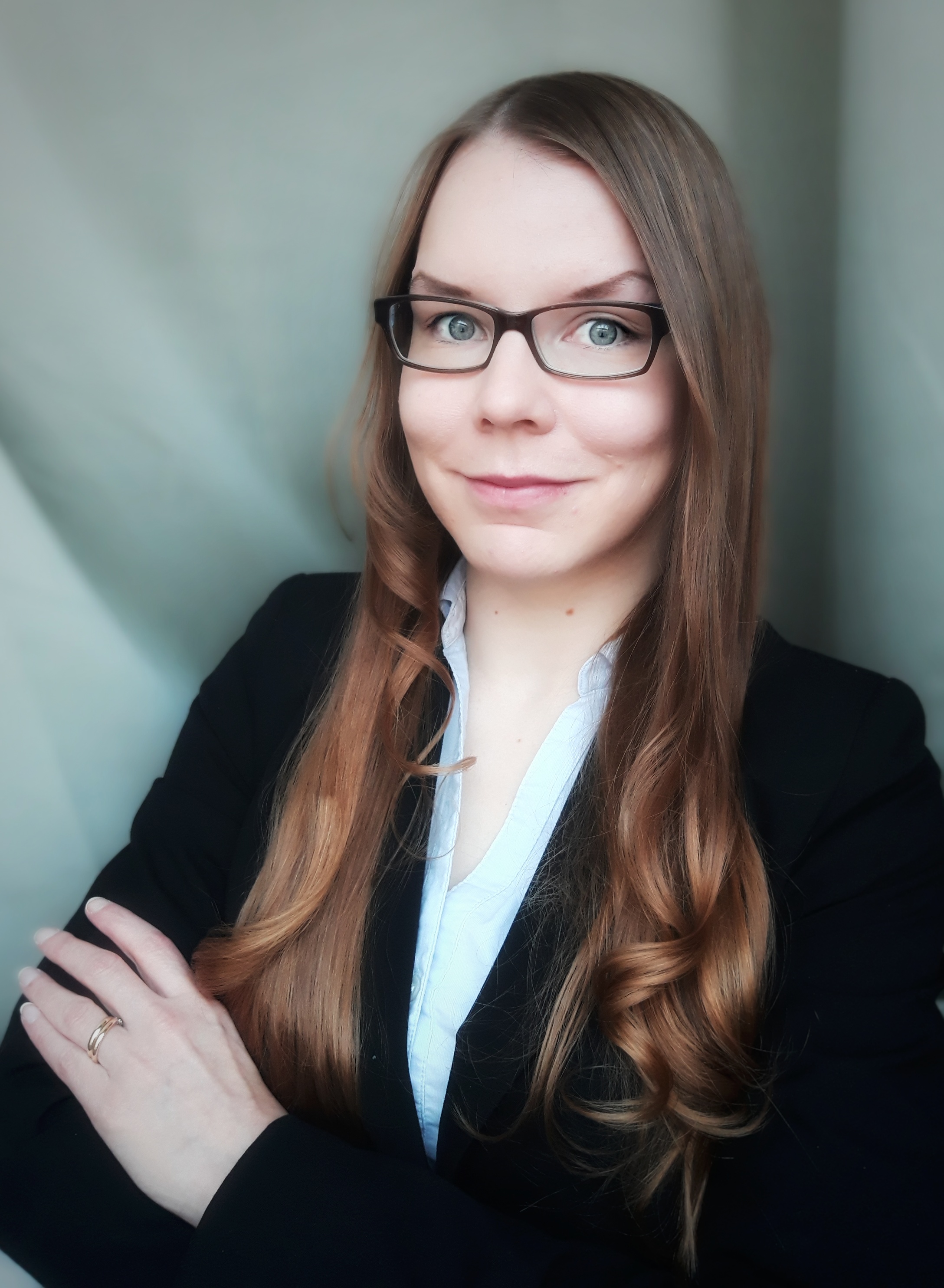 Henkilö pukeutunut mustaan jakkuun ja vaaleansiniseen paitaan, pitkät vaaleanruskeat hiukset ja silmälasit, katsoo kameraan. Hänen kätensä ovat ristissä rinnalla.