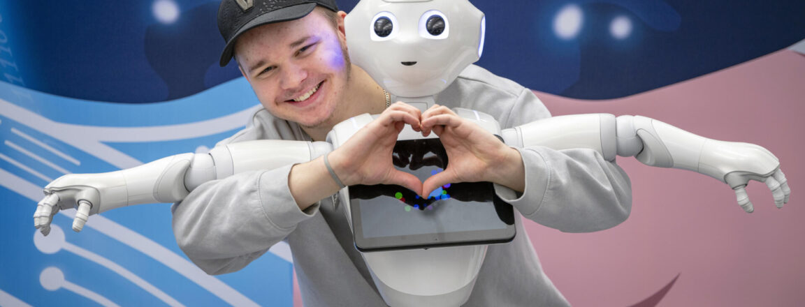 Iloinen henkilö seisoo robotin takana ja muodostaa käsillään sydämen. Robotti näyttää humanoidilta ja sillä on näyttö rinnassa. Taustalla on futuristinen kuviointi.