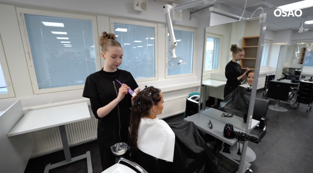 Hiusalan opiskelija värjää asiakkaan hiuksia kampaamossa. Asiakas istuu peilin edessä ja opiskelija levittää hiusväriä siveltimellä.