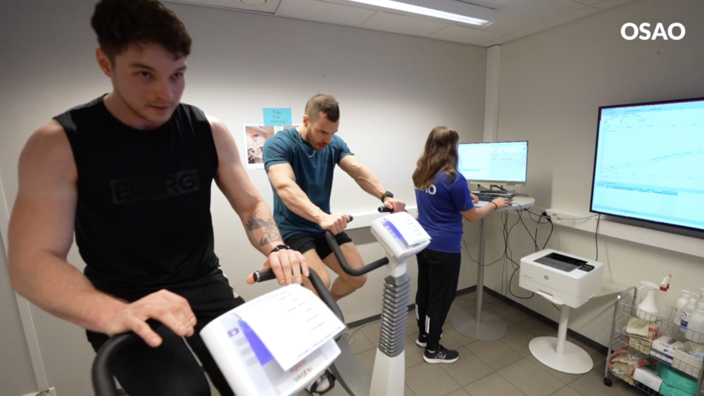 Kaksi miestä polkee kuntopyörää laboratoriossa ja nainen seuraa heidän suoritustaan tietokoneella. Taustalla näkyy suuria näyttöjä, joissa on graafisia esityksiä.