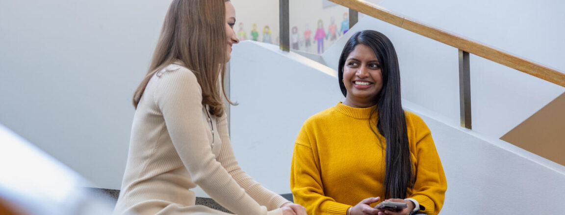 Kaksi naista keskustelee portailla. Vasemmalla on vaaleahiuksinen nainen beige mekossa ja oikealla nainen keltaisessa neuleessa, joka pitää puhelinta käsissään. Taustalla näkyy valkoiset seinät ja kaide.