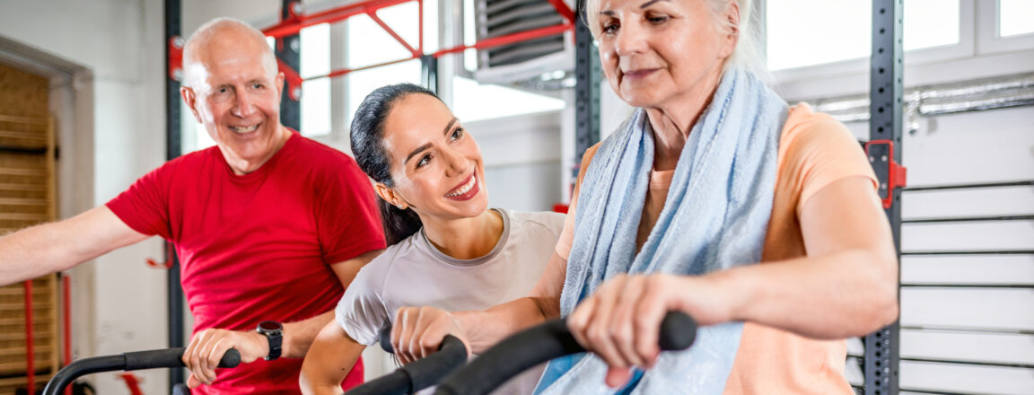 Kolme henkilöä kuntosalilla, kaksi vanhempaa aikuista harjoittelee istuen kuntopyörillä, ja nuorempi henkilö hymyilee ja kannustaa heitä.
