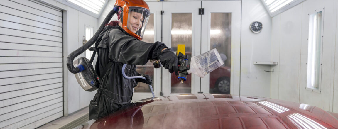Henkilö maalaa auton osaa suihkumaalilla autotallissa tai työpajassa, yllään suojavarusteet kuten oranssi kypärä ja hengityssuojain. Auton osa on kiiltävä ja punainen.