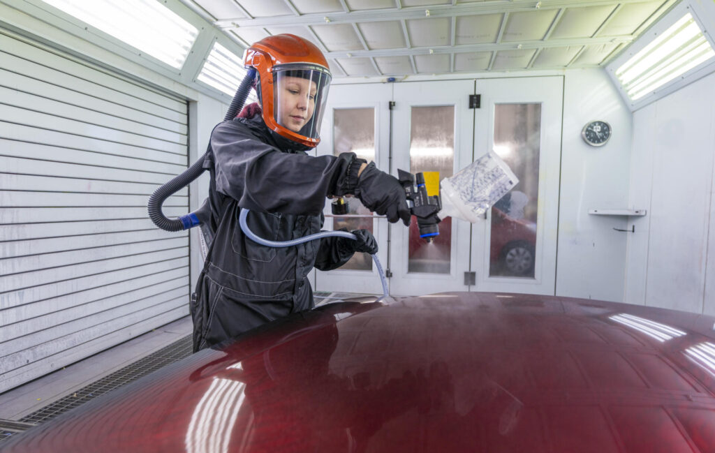 Henkilö maalaa auton konepeltiä maalauskopissa suojavarusteisiin pukeutuneena. Hänellä on kypärä ja hengityssuojain, ja hän käyttää maalisuihkupistoolia.