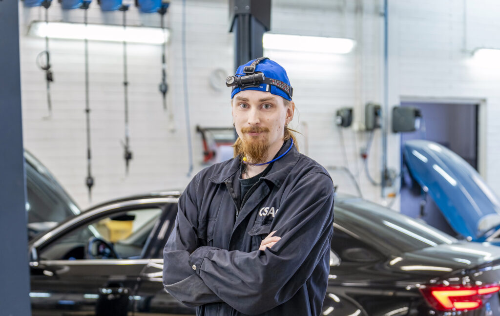 Diagnoosimekaanikko seisoo korjaamossa ristissä käsin, päässään sininen lakki ja otsalamppu. Taustalla on musta auto.