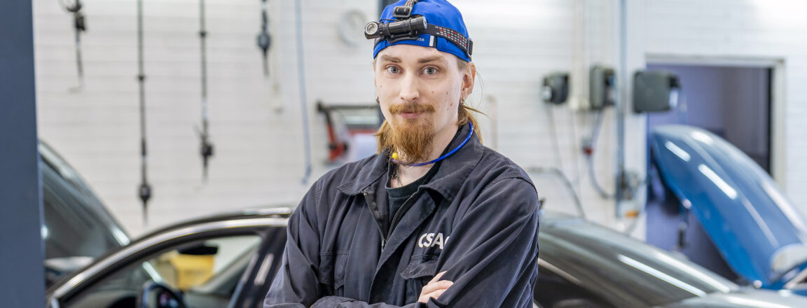 Diagnoosimekaanikko seisoo korjaamossa ristissä käsin, päässään sininen lakki ja otsalamppu. Taustalla on musta auto.