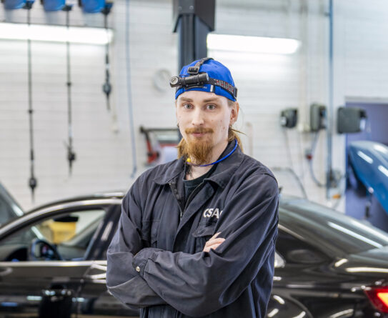 Diagnoosimekaanikko seisoo korjaamossa ristissä käsin, päässään sininen lakki ja otsalamppu. Taustalla on musta auto.