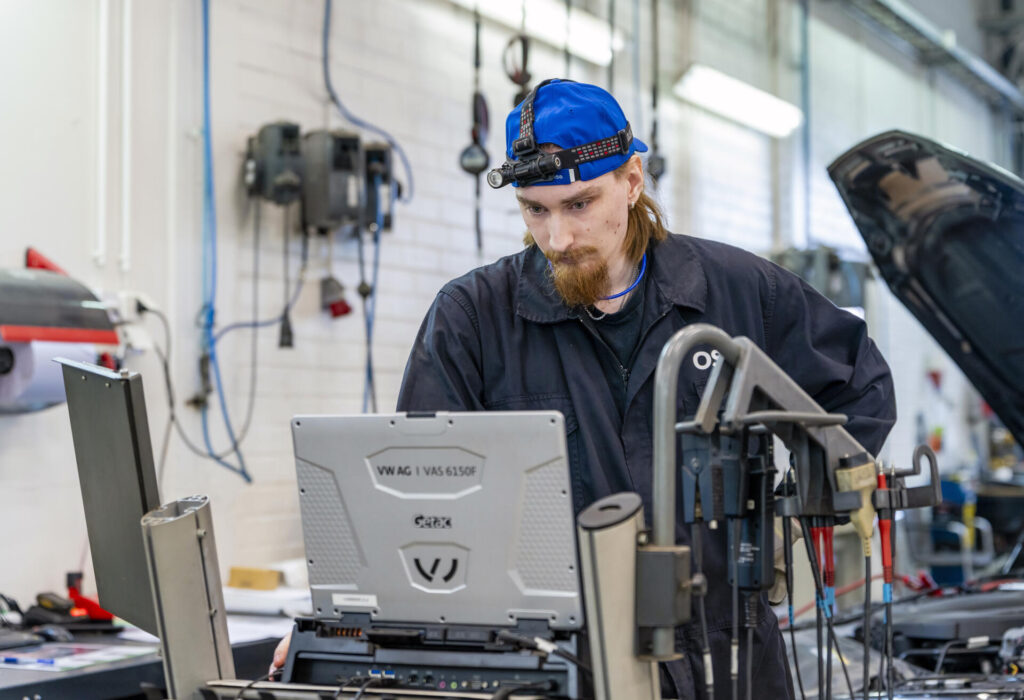 Mekaanikko työskentelee autokorjaamolla kannettavan tietokoneen kanssa. Hänellä on päällään sininen lippalakki ja otsalamppu. Taustalla on auton konepelti auki.