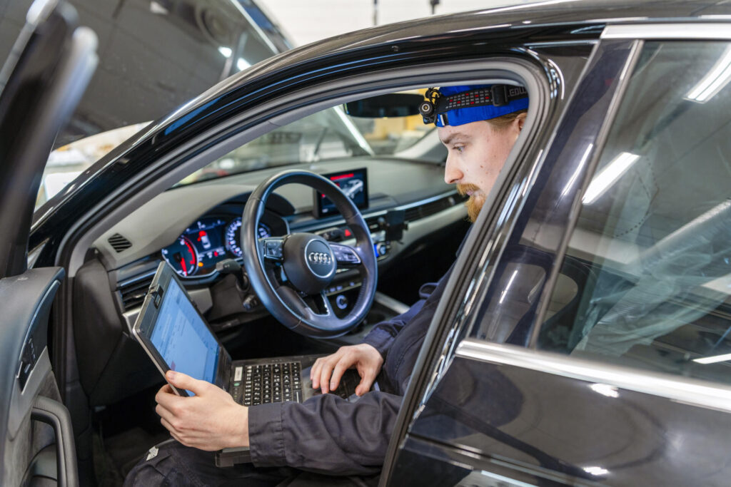 Henkilö istuu auton etuistuimella kannettavan tietokoneen kanssa. Hänellä on otsalamppu päässään ja hän tekee diagnostiikkatarkastusta ajoneuvolle. Auto on sisätiloissa, ja kojelaudan valot ovat päällä.