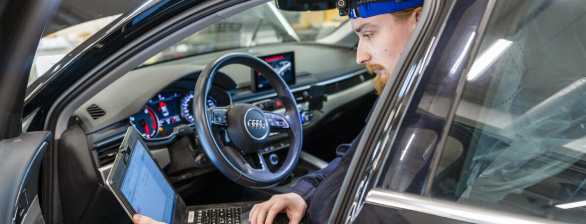 Henkilö istuu auton etuistuimella kannettavan tietokoneen kanssa. Hänellä on otsalamppu päässään ja hän tekee diagnostiikkatarkastusta ajoneuvolle. Auto on sisätiloissa, ja kojelaudan valot ovat päällä.