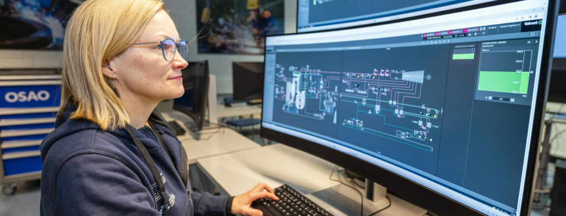 Nainen istuu työpöydän ääressä tietokoneen äärellä, jossa on kaksi suurta näyttöä, joilla näkyy teknisiä kaavioita. Hänellä on silmälasit ja hän käyttää näppäimistöä. Taustalla on teknisiä kuvia seinällä.