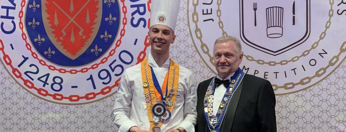 Kaksi miestä seisoo yhdessä palkintojenjakotilaisuudessa. Vasemmalla oleva henkilö on pukeutunut valkoiseen kokin takkiin, kokin hattuun ja kantaa koristeellista nauhaa kaulassaan, ja hänellä on palkinto käsissään. Oikealla oleva henkilö on pukeutunut smokkiin ja kantaa myös koristeellista nauhaa kaulassaan. Taustalla näkyy kaksi suurta kilpeä, joissa lukee 'Chaîne des Rôtisseurs' ja 'Jeunes Chefs Rôtisseurs Competition'.