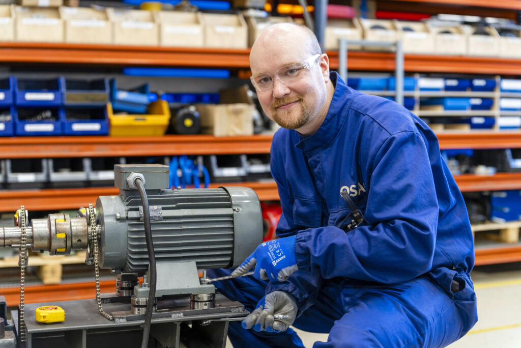 Mies sinisessä suojahaalarissa ja suojalaseissa työskentelee teollisuusympäristössä. Hän polvistuu moottorin viereen ja osoittaa siihen hymyillen. Taustalla on hyllyjä, joissa on työkaluja ja tarvikkeita.